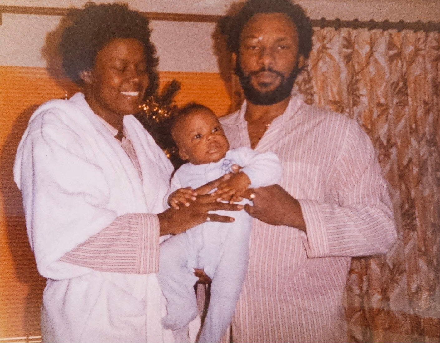 Family of three indoors, with a black woman holding a baby and a black man standing next to them. Black family
