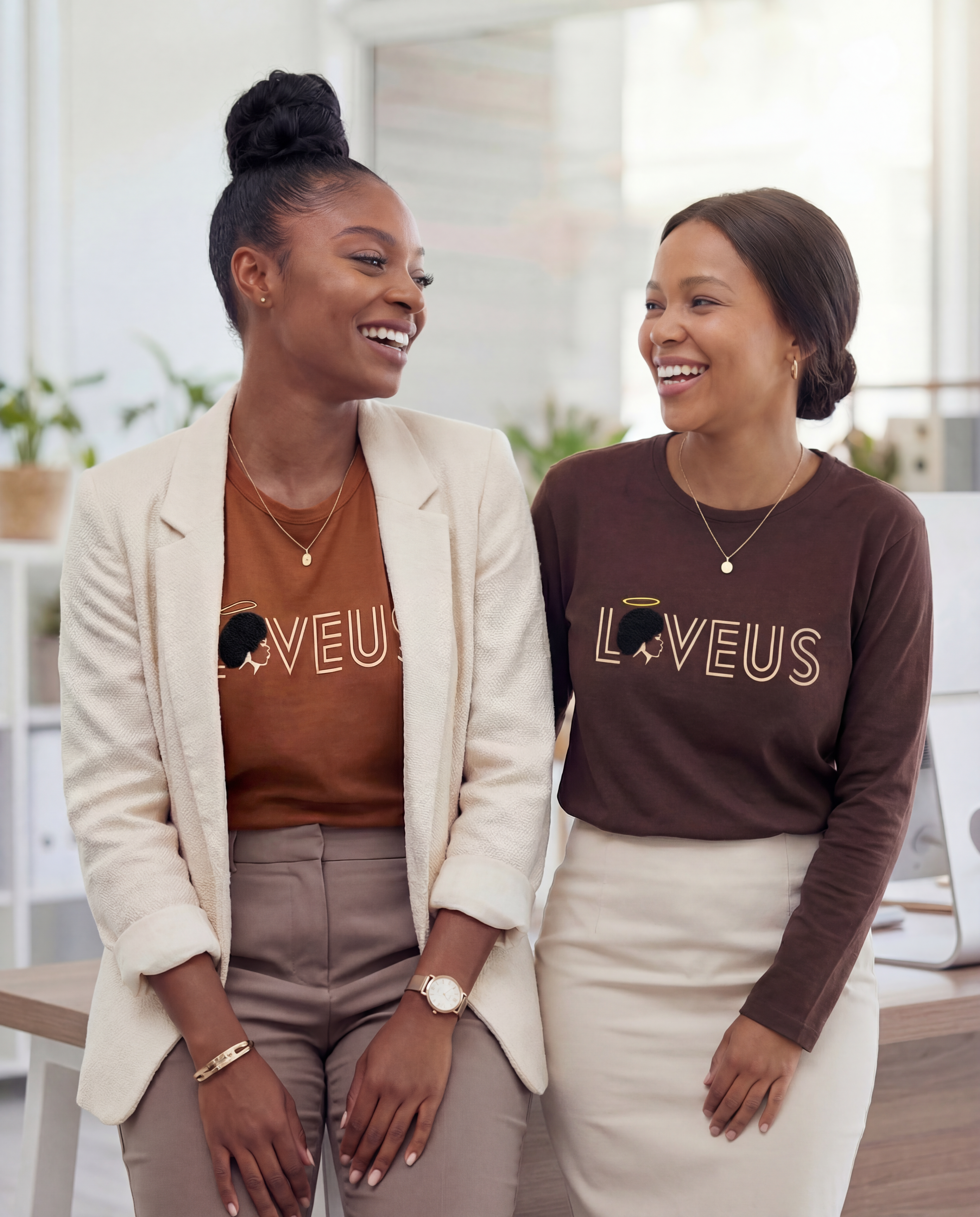 Two women wearing matching 'LOVEUS' shirts in an office setting. Caramel Angel and Chocolate Angel cotton shirt in warm earth tone with raised embroidered Afro design mimicking natural hair texture, relaxed structured fit LOVEUS. Office setting 
