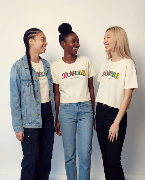 Three women wearing 'LOVEUS' t-shirts standing together against a plain background. Extremely Gifted cropped cotton shirt with abstract graphic design, relaxed fit with dropped shoulders and shorter length LOVEUS.