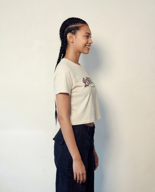 Person wearing a white t-shirt with colorful text and dark pants against a plain background. Extremely Gifted cropped cotton shirt with abstract graphic design, relaxed fit with dropped shoulders and shorter length LOVEUS