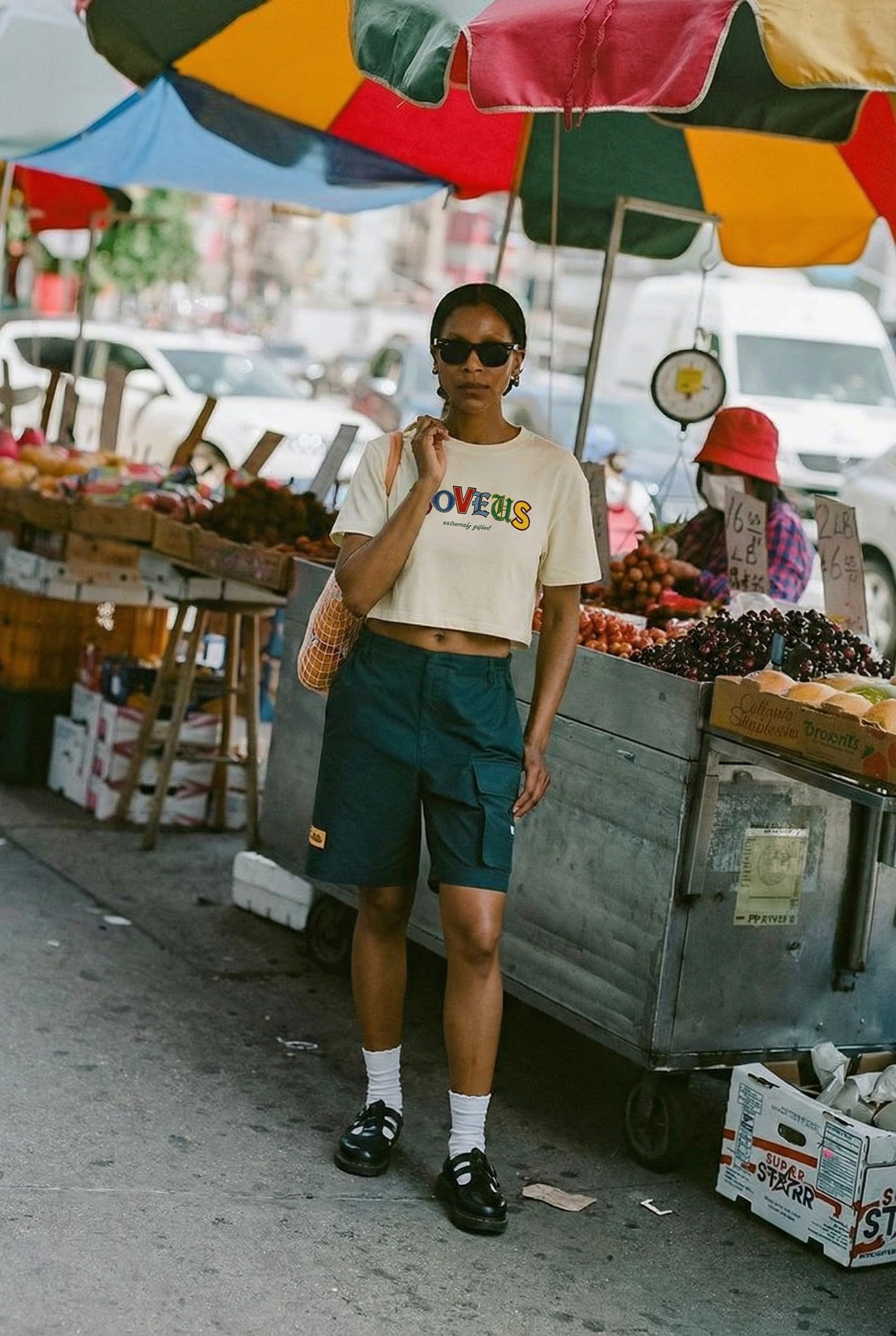 Woman standing in an outdoor fruit market setting with colorful umbrellas and fruit stands. Extremely Gifted cropped cotton shirt with abstract graphic design, relaxed fit with dropped shoulders and shorter length LOVEUS. Streetwear shot.